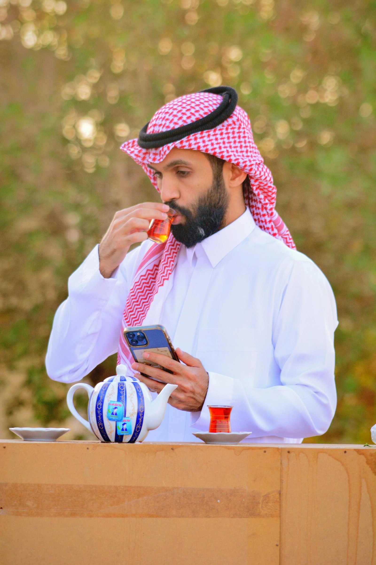 Man wearing keffiyeh drinking tea outdoors, using cellphone in Hafar Al Batin.